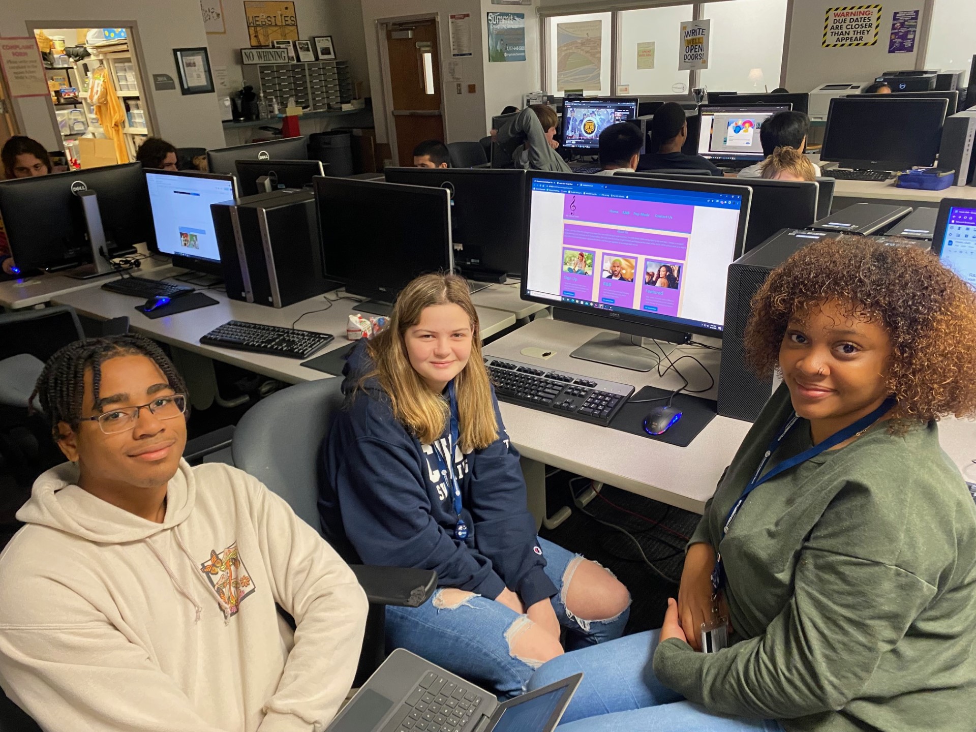 Group of students, sitting in their own grey rolling chairs at a long, white table with a set of computers, keyboards, and mice in front of each student.